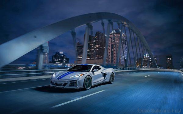 Front 3/4 view of 2024 Chevrolet Corvette E-Ray 3LZ convertible in Silver Flare with Electric Blue stripe package driving across a city bridge at night. Pre-production model shown. Actual production model may vary. Model year 2024 Corvette E-Ray available 2023.